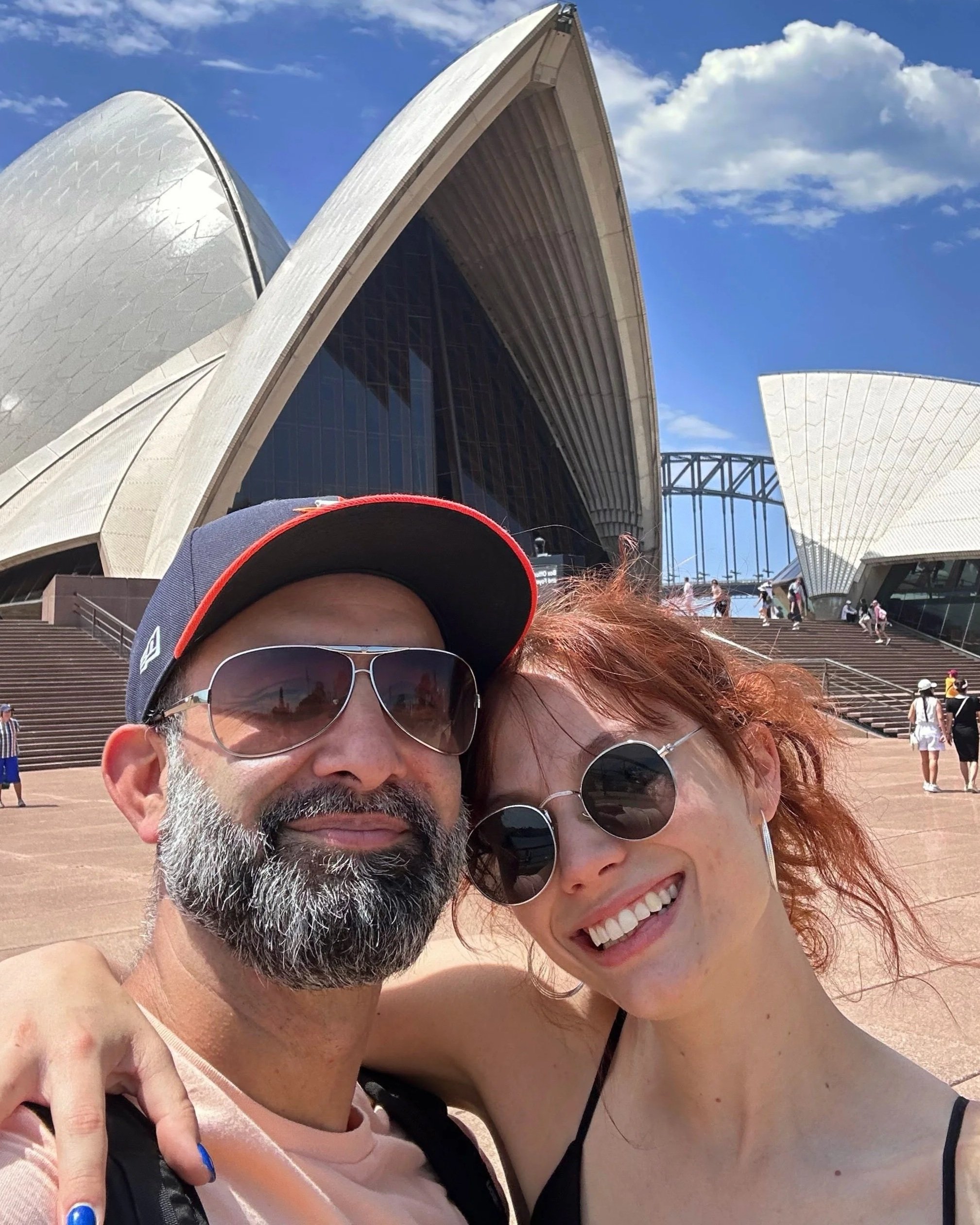 Close-up of a smiling man with a beard and sunglasses, and a smiling woman with curly red hair and sunglasses, taking a selfie in front of the Sydney Opera House, with steps and tourists in the background.