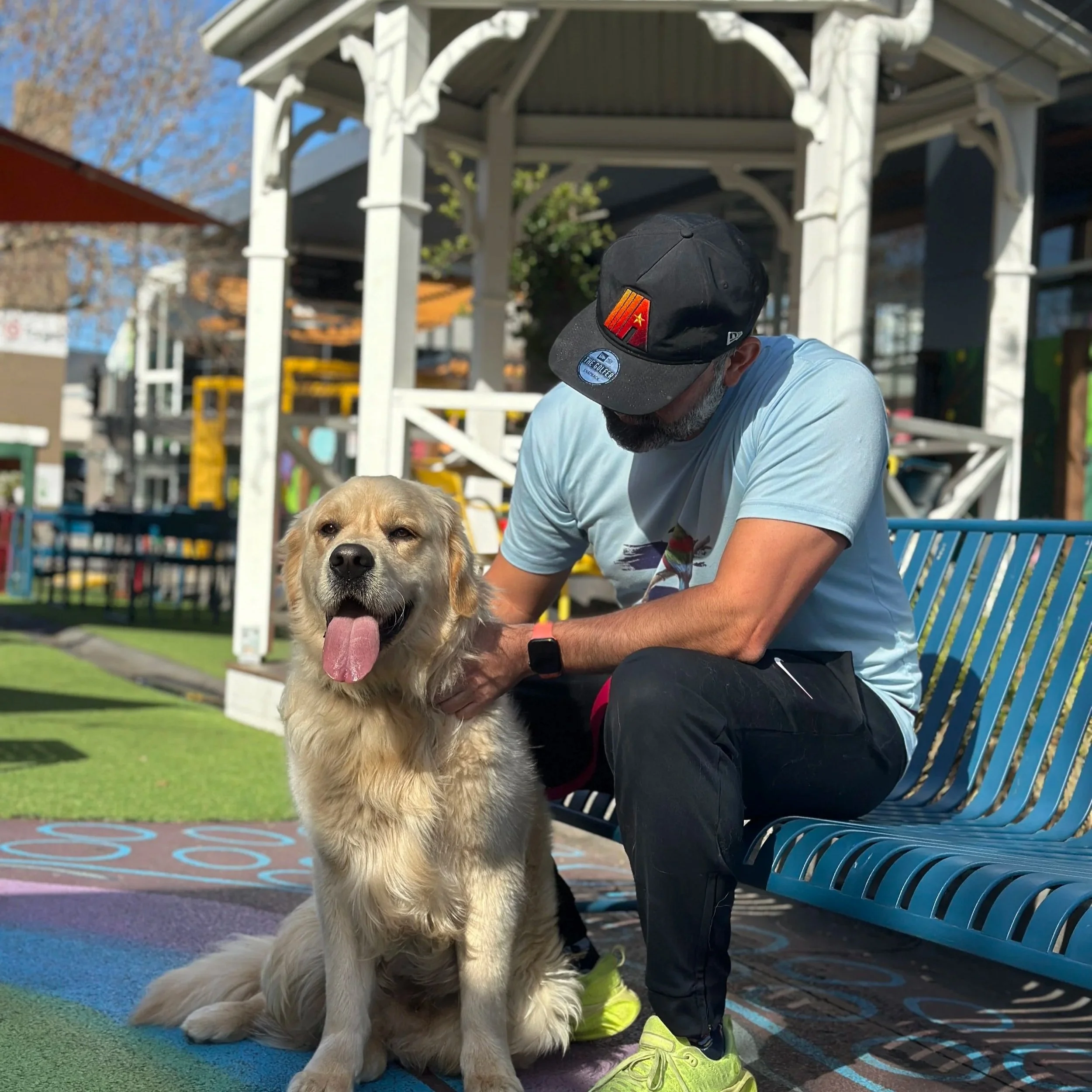 A golden retriever dog with its tongue out sitting on a colorful playground surface, next to a person wearing a black cap and neon green shoes, under a white pavilion with a boy in a blue shirt.