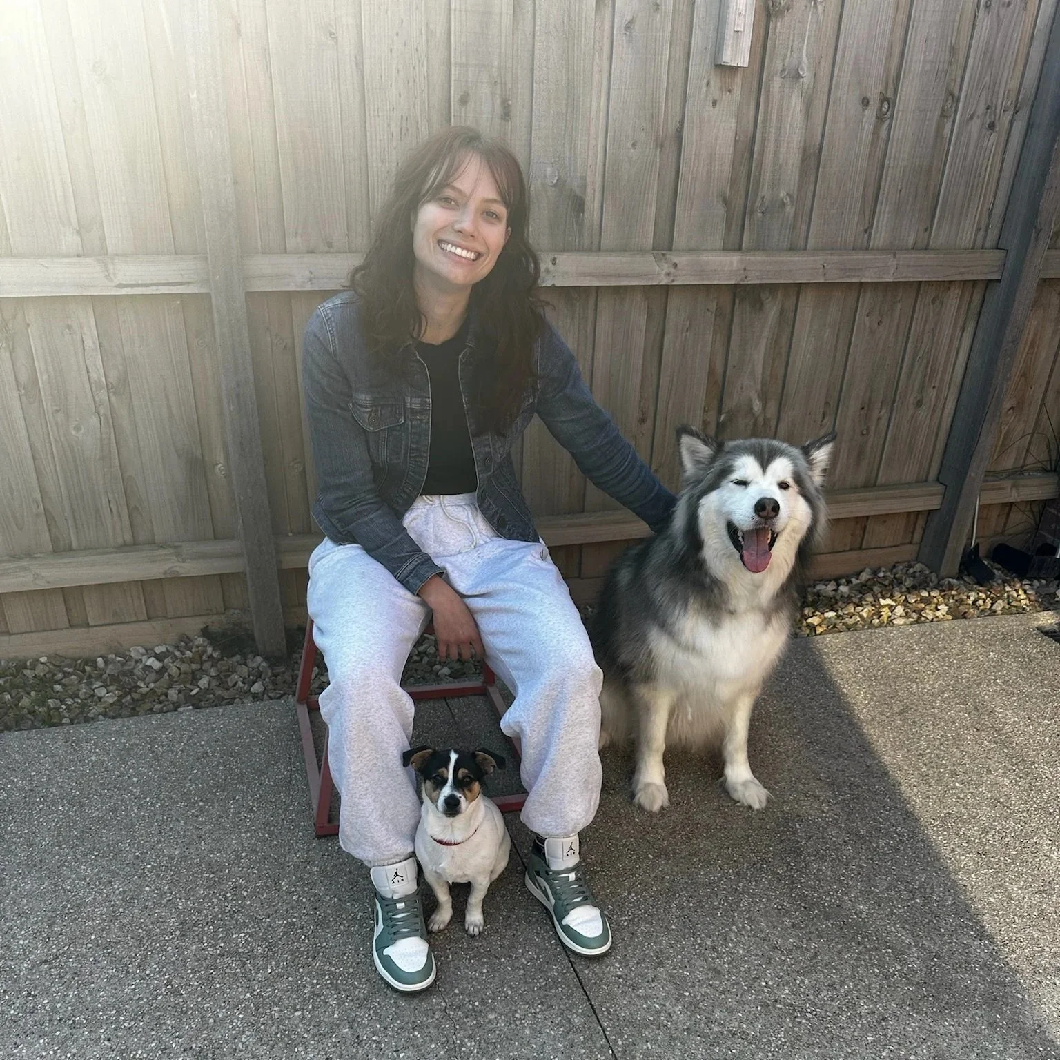 A woman with dark wavy hair, wearing a denim jacket and white sweatpants, sitting on a small red stool, smiling with her hand on a large, fluffy Siberian Husky. A small, black and white dog sits on her lap. They are outside in front of a wooden fence on a concrete surface.