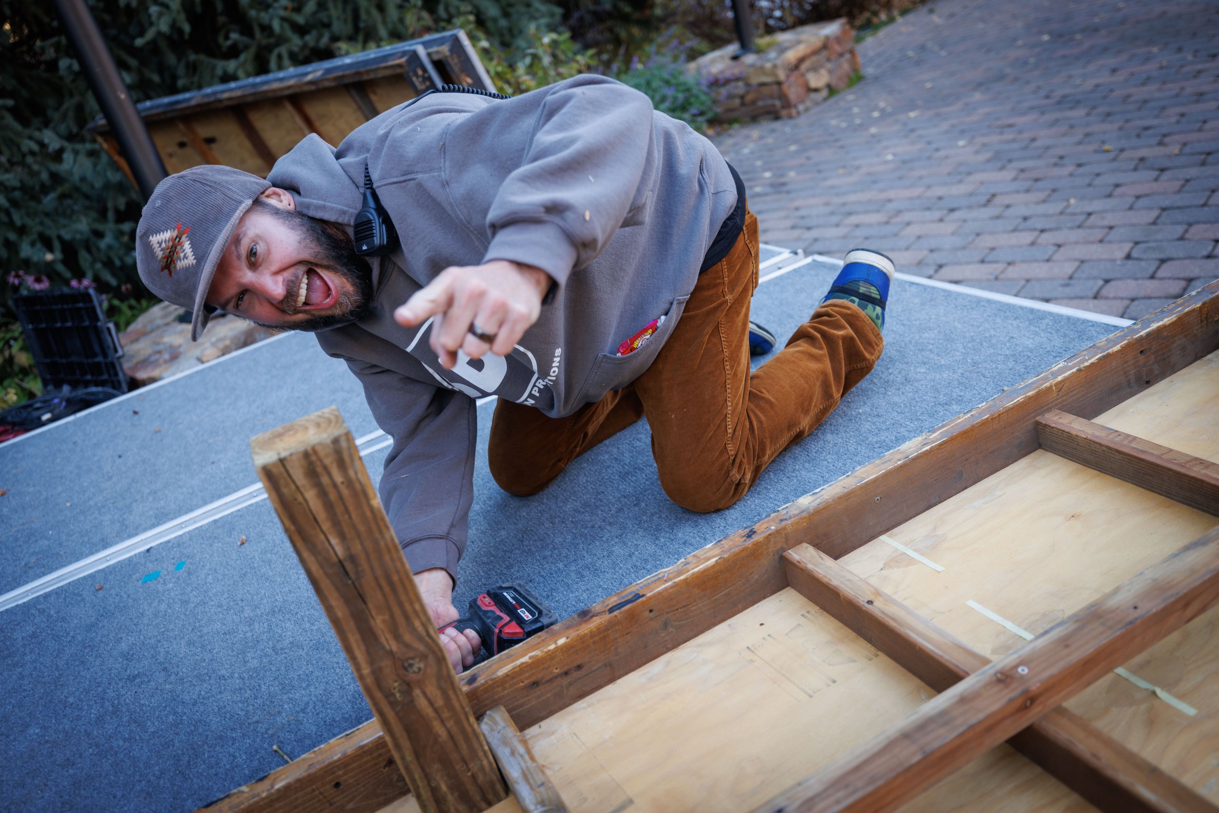 A man with a beard wearing a gray hoodie, brown pants, and a gray beanie with a holiday pattern is kneeling on a wooden frame, smiling and pointing at the camera, working on a woodworking project outdoors on a brick patio.