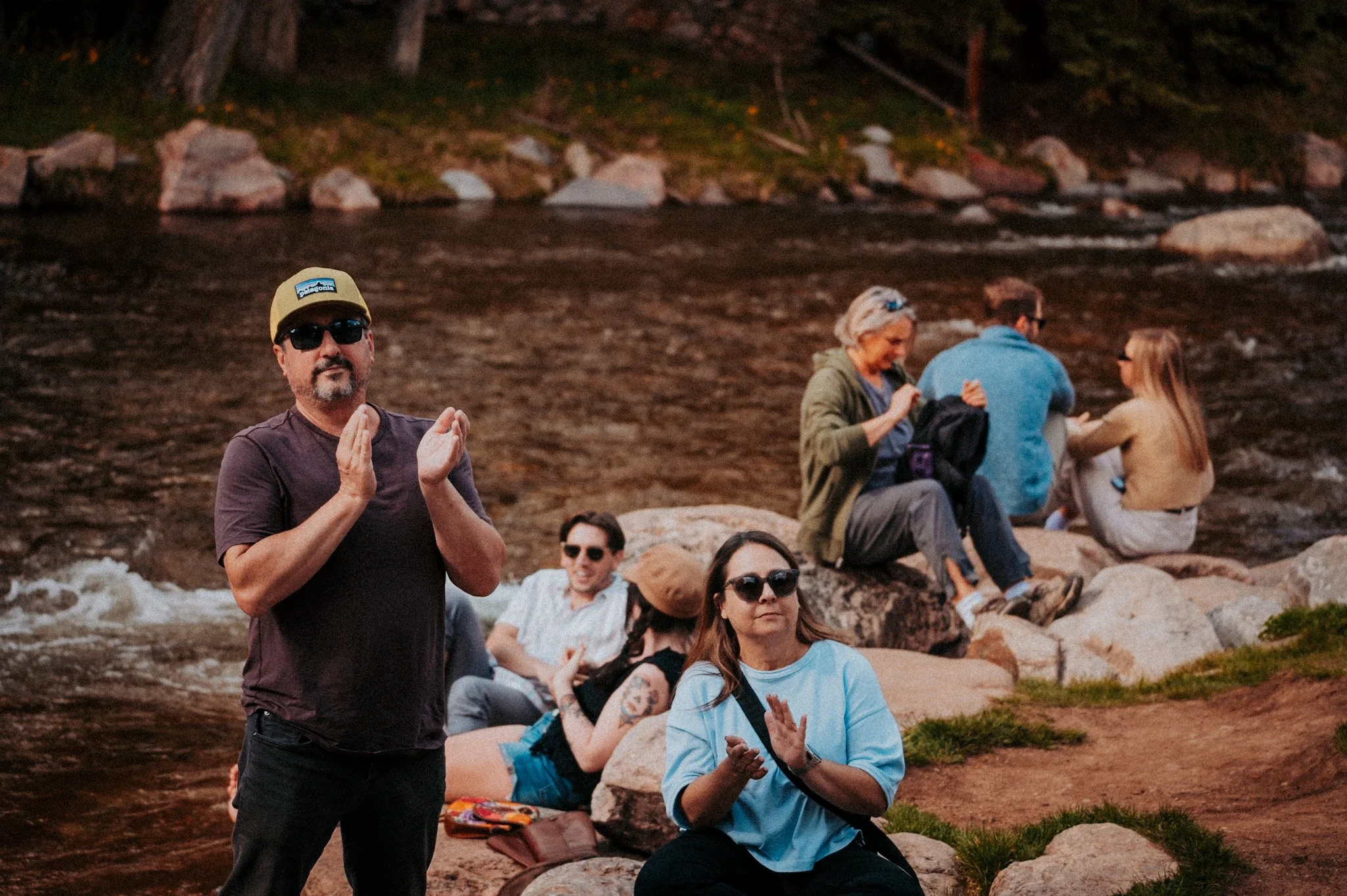 A group of people sitting and standing on rocks by a river at sunset, some clapping, others talking and relaxing.