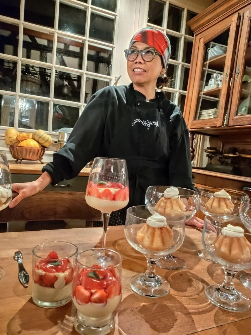 A woman wearing glasses, a red headband, and a black apron stands behind a wooden kitchen counter with desserts. The desserts include strawberry parfaits in glasses, topped with whipped cream, and panna cotta or similar desserts in cocktail glasses with a fluted shape, topped with whipped cream. There is a bowl of squash or gourds and a window with multiple small panes in the background.