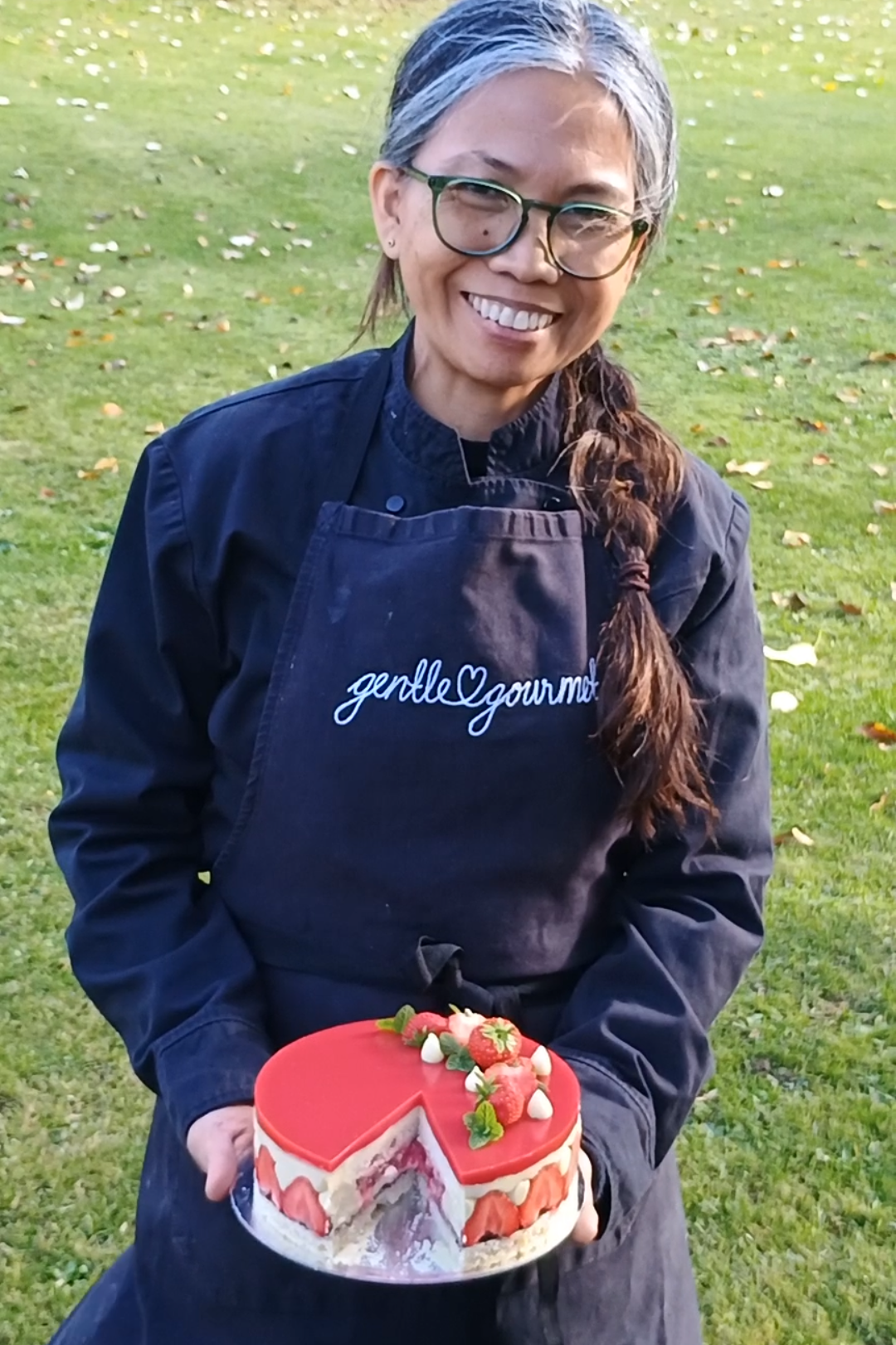 A woman wearing glasses and a black apron with 'gentleGourmet' embroidered on it, smiling and holding a decorated strawberry cake outdoors on a grassy field. The Gentle Gourmet Institute Normandy France. Hayden's House Food and Produce