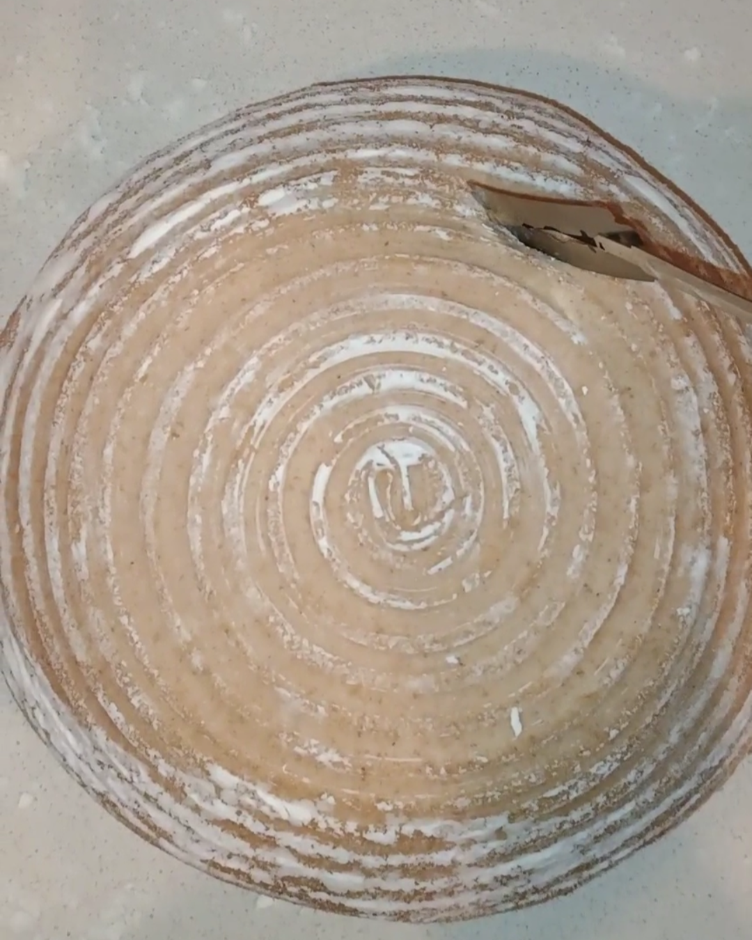 A round wooden cutting board with concentric circles, dusted with flour, and a metal spatula resting at the edge. Baking sourdough at Hayden's House Food and Produce