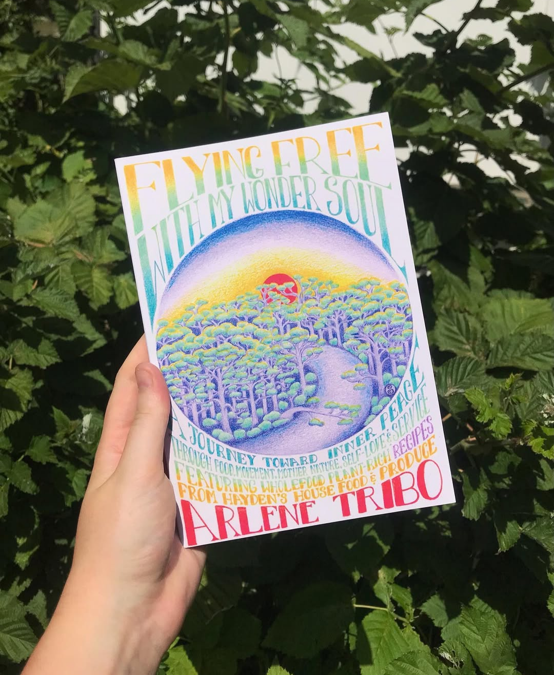 A colorful book cover held in a hand against green foliage. The cover features an illustration of a landscape with trees, a hill, and a red sun, with pastel shades of yellow, purple, and green. The title reads 'Flying Free with My Wonder Soul' and the author is Arlene Tribo.