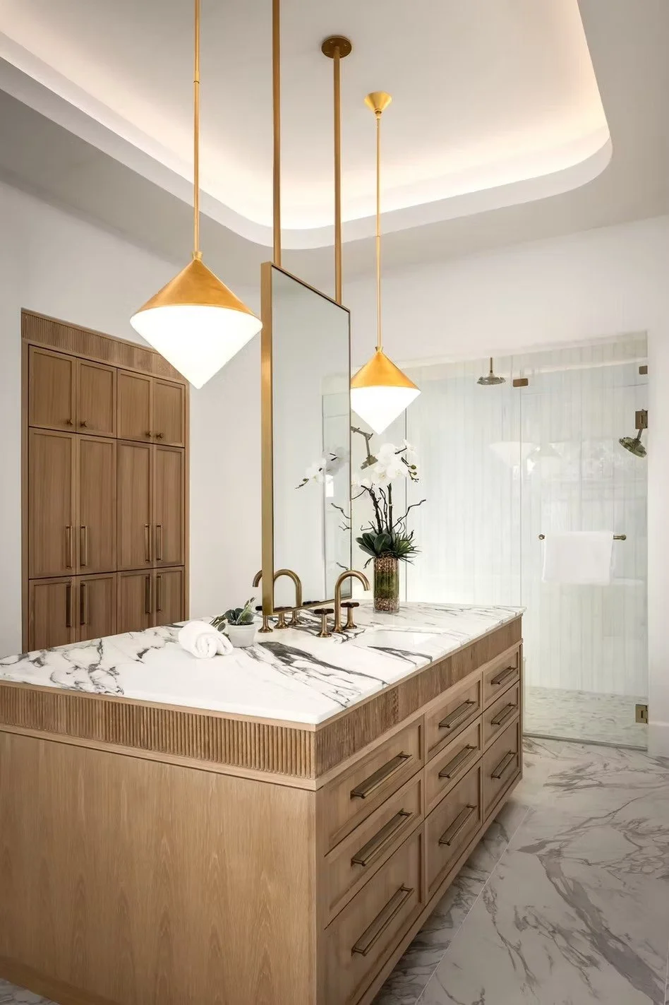 Modern bathroom with marble countertop, wooden cabinetry, and gold fixtures, including pendant lights and a faucet, with a glass shower door and a decorative plant.