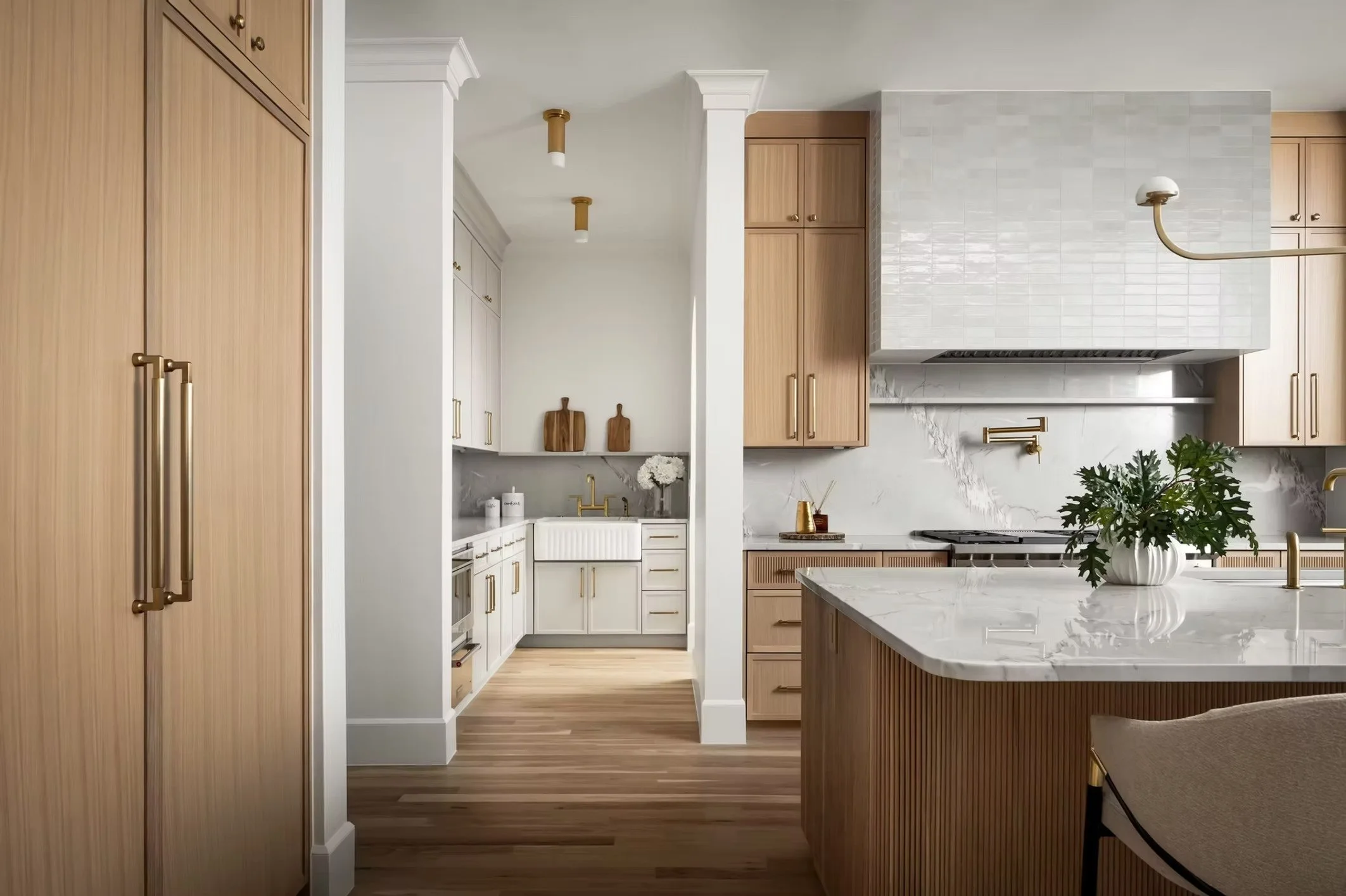 Modern kitchen with light wood cabinets, white marble countertops, and a white tiled backsplash. Gold fixtures and hardware, and green plants on the counter.
