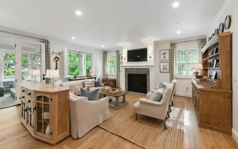 A cozy living room with beige sofas, a brick fireplace, wooden furniture, and large windows showing greenery outside.
