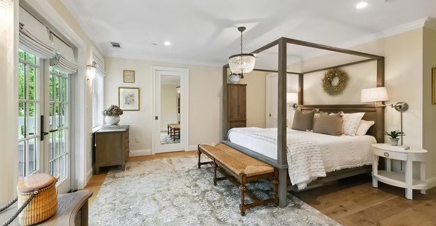 Bright bedroom with a four-poster bed, nightstand with lamp, wicker bench at the foot, and sliding glass doors with curtains leading outside.