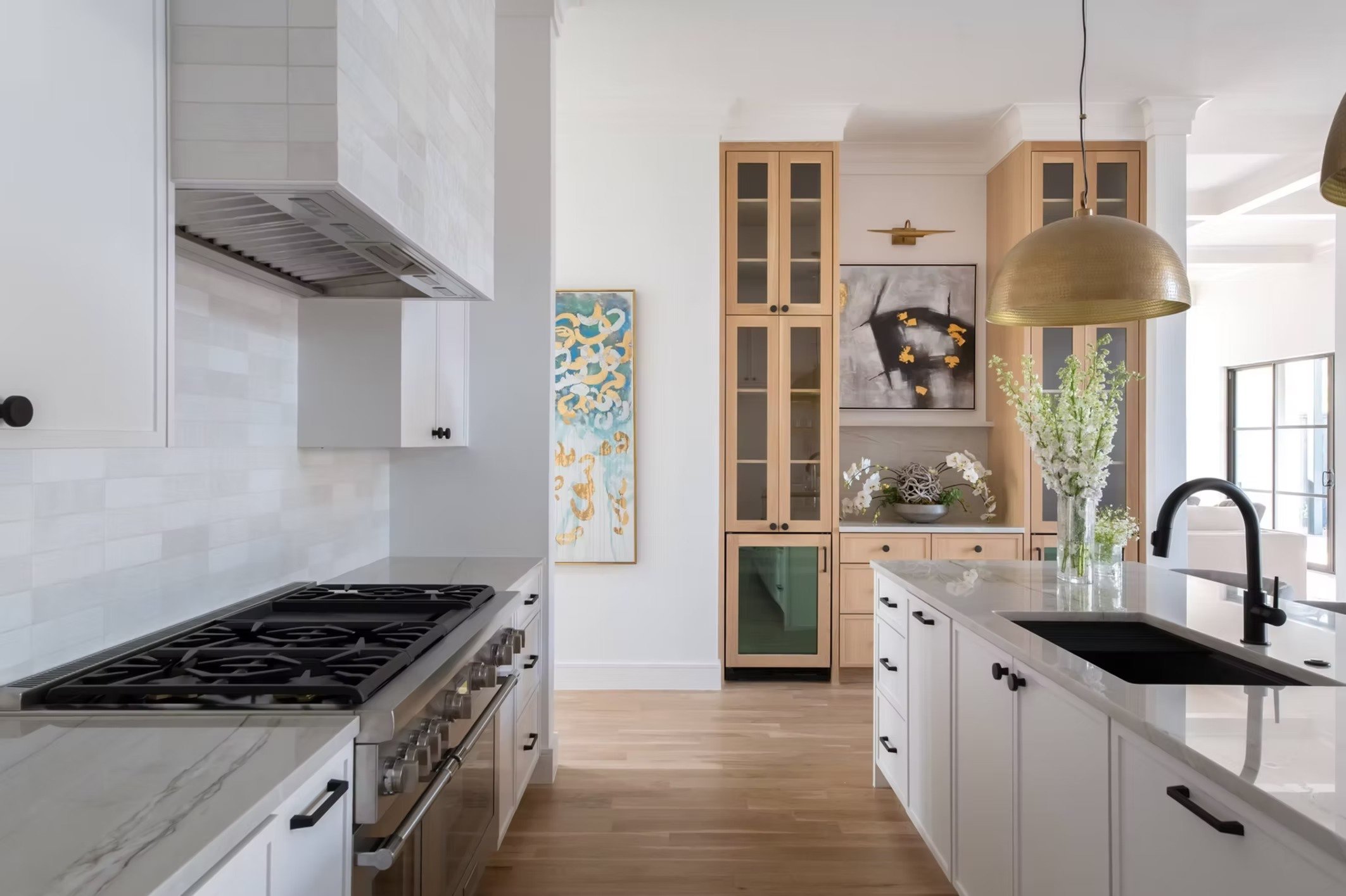 Modern kitchen with white cabinets, a marble countertop, a black sink with a gooseneck faucet, and a hanging gold light fixture. There are wooden cabinets with glass doors, artwork, and a vase with white flowers.