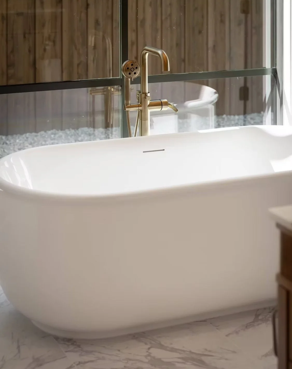 White freestanding bathtub with a gold faucet and a handheld shower, placed in a bathroom with a glass wall, wooden paneling, and marble floor.