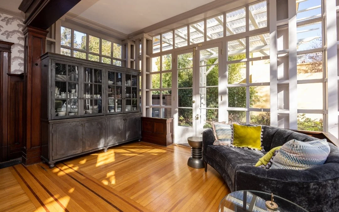 Living room with large windows, a black velvet sofa with colorful pillows, a wooden cabinet, and hardwood floors.