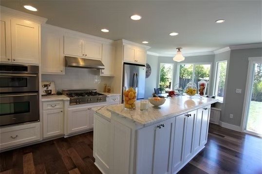 Modern kitchen with white cabinets, a large marble island, stainless steel appliances, hardwood floors, and a bright sunlit breakfast nook.