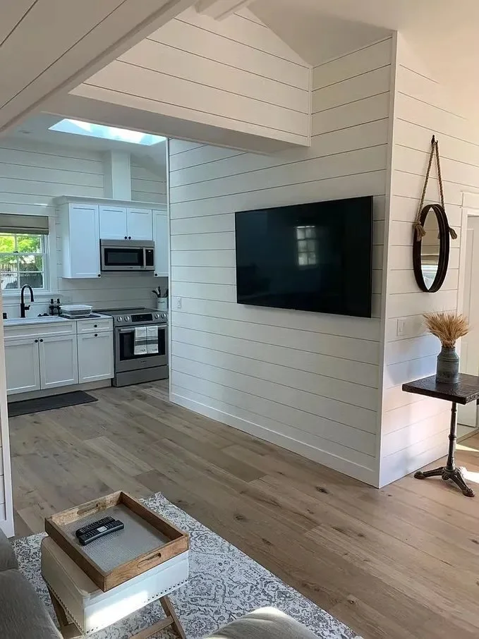 Modern kitchen with white cabinets, stainless steel appliances, and a living area with a wall-mounted flat-screen TV, white shiplap walls, and wooden flooring.