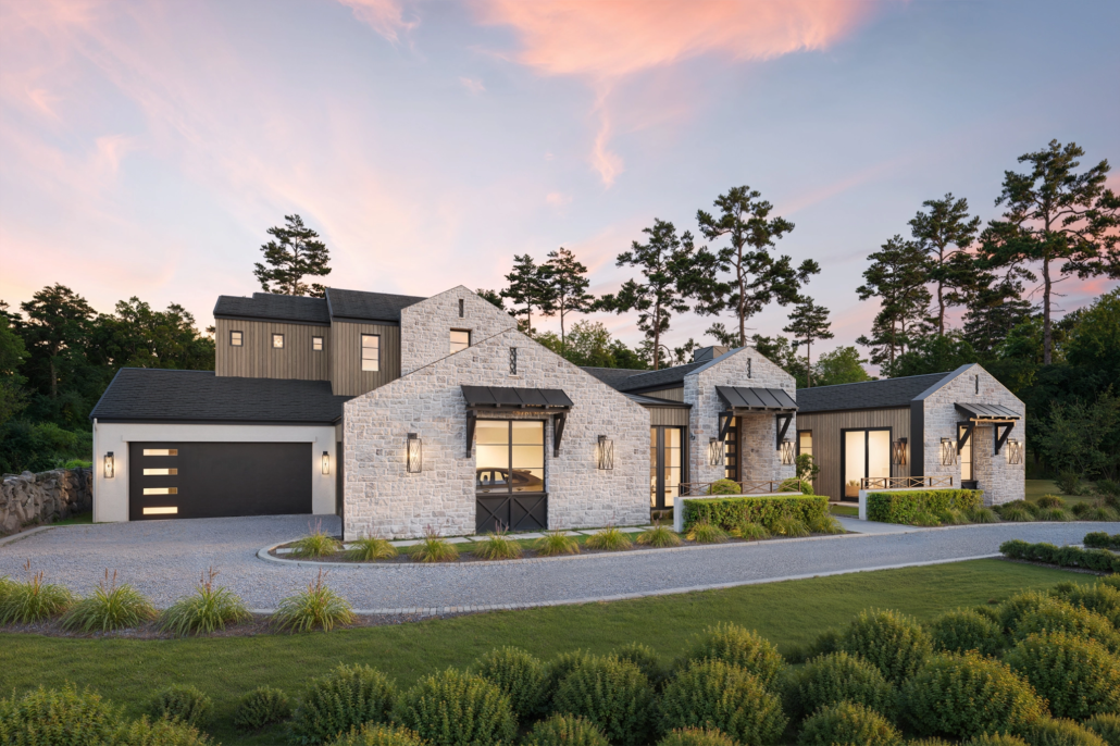 Modern farmhouse with white stone and dark wood exterior, large windows, and a gravel driveway at sunset.