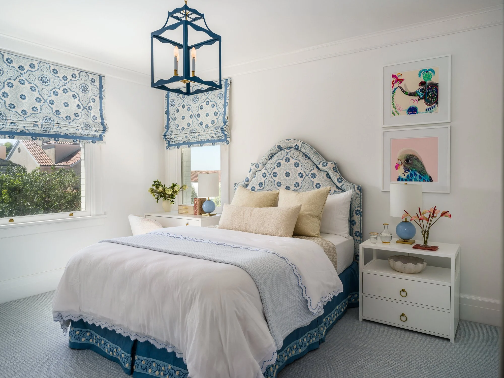 Bright bedroom with white walls and a window with blue and white patterned Roman shades. A bed with white and beige pillows, a white dresser with books, a vase with flowers, and a lamp. Decor includes framed colorful animal art on the wall.