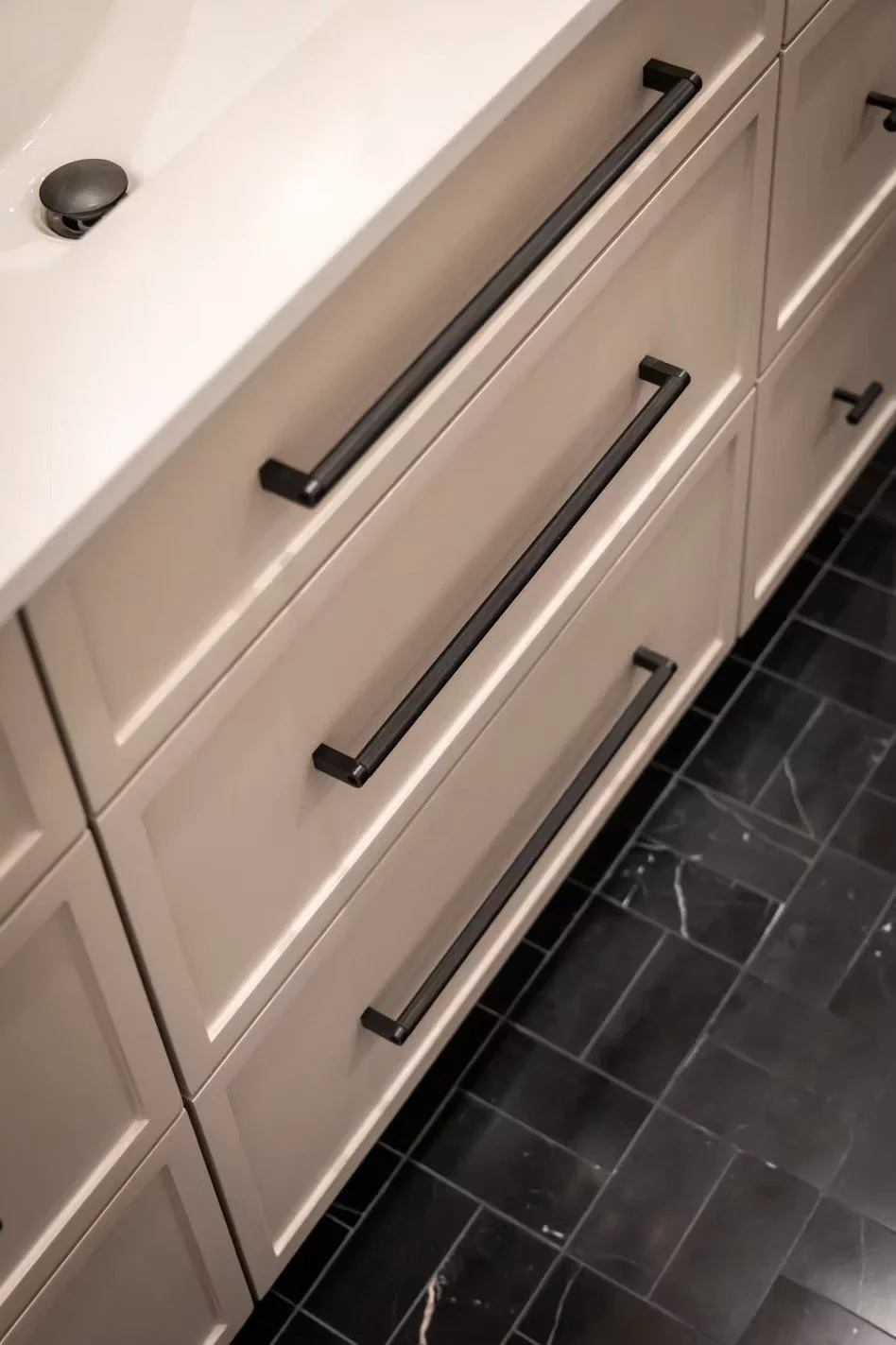 A bathroom vanity with four beige drawers featuring long black handles, and a white countertop with a black drain stopper and plumbing fixtures.