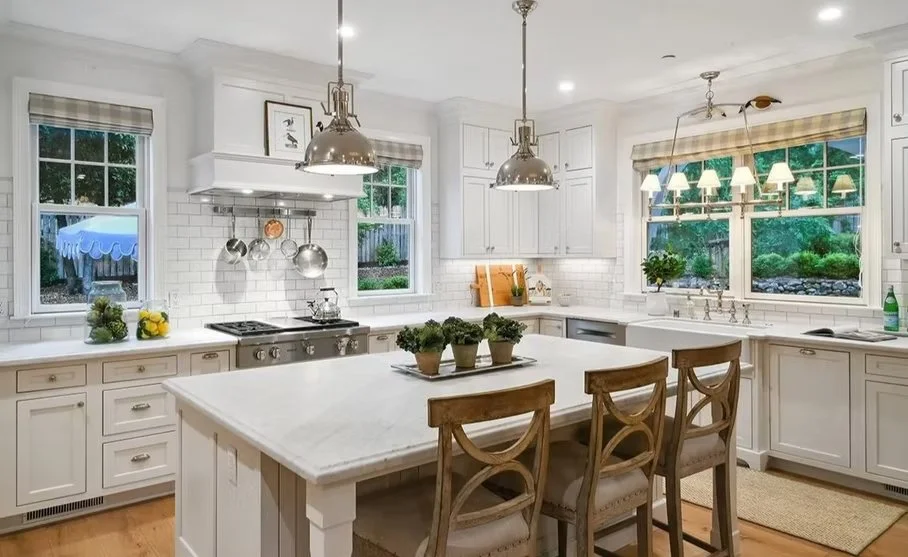 Bright white kitchen with island, hanging pendant lights, window with plaid valance, potted plants on windowsill, and a kitchen stove with utensils hanging above.