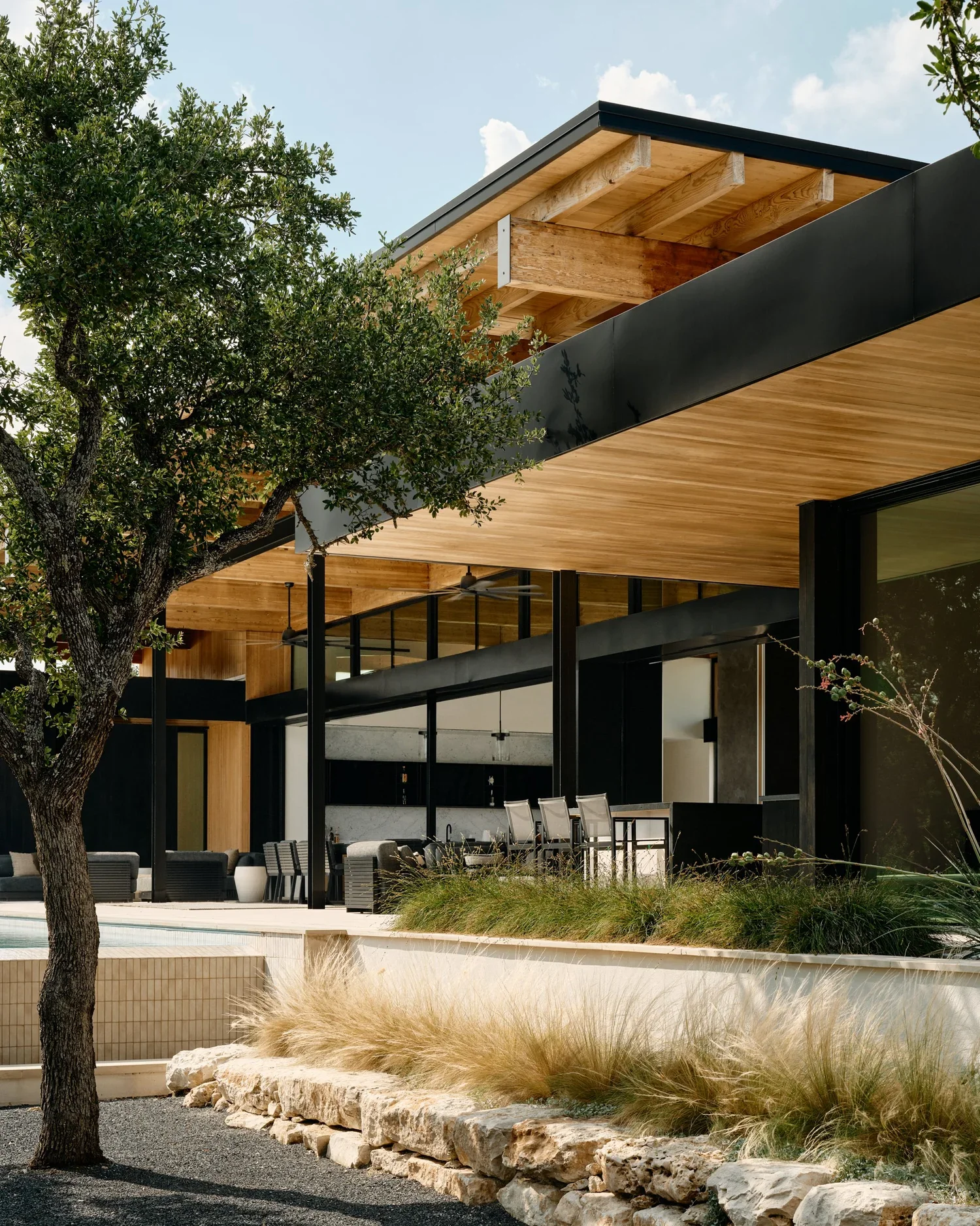 Modern house with wooden and black exterior design, outdoor furniture, palm trees, and landscaped garden with rocks and grasses.
