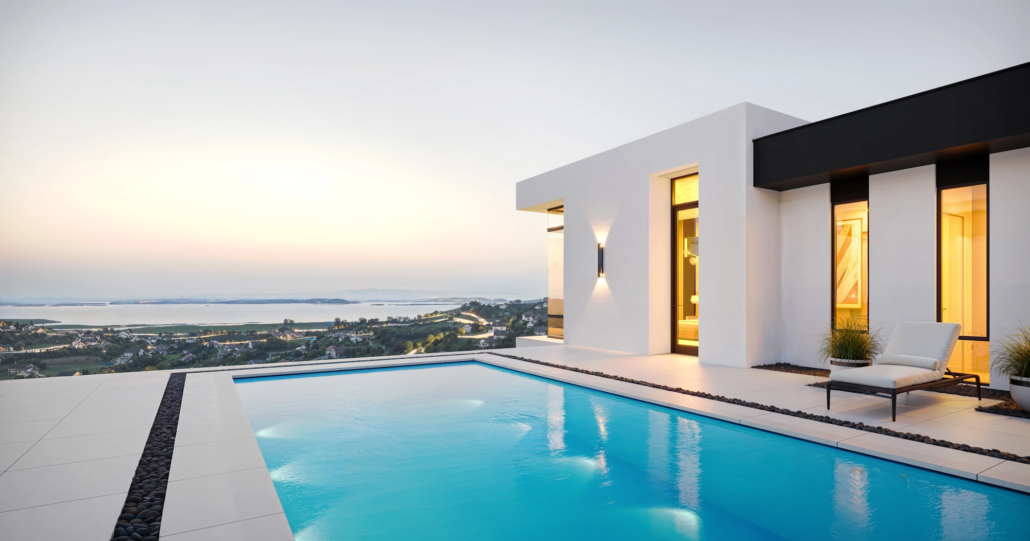 Modern white house with large swimmer pool and outdoor lounge chair on a patio at sunset, overlooking a cityscape.