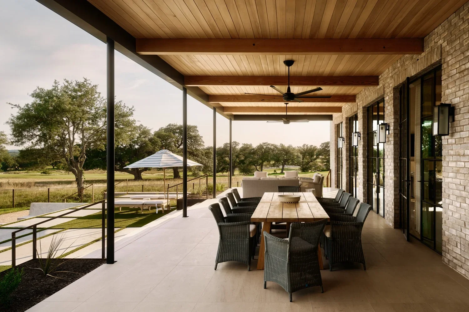 Covered patio with a wooden ceiling, ceiling fans, and black wall-mounted lights, overlooking a grassy yard with trees and an outside umbrella with seating area.