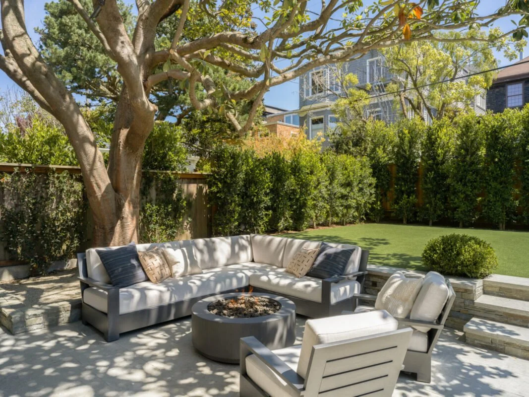 Outdoor patio with a large tree providing shade, a white sectional sofa with pillows, two armchairs, a round fire pit, and a lush green yard enclosed by a wooden fence and tall hedge.