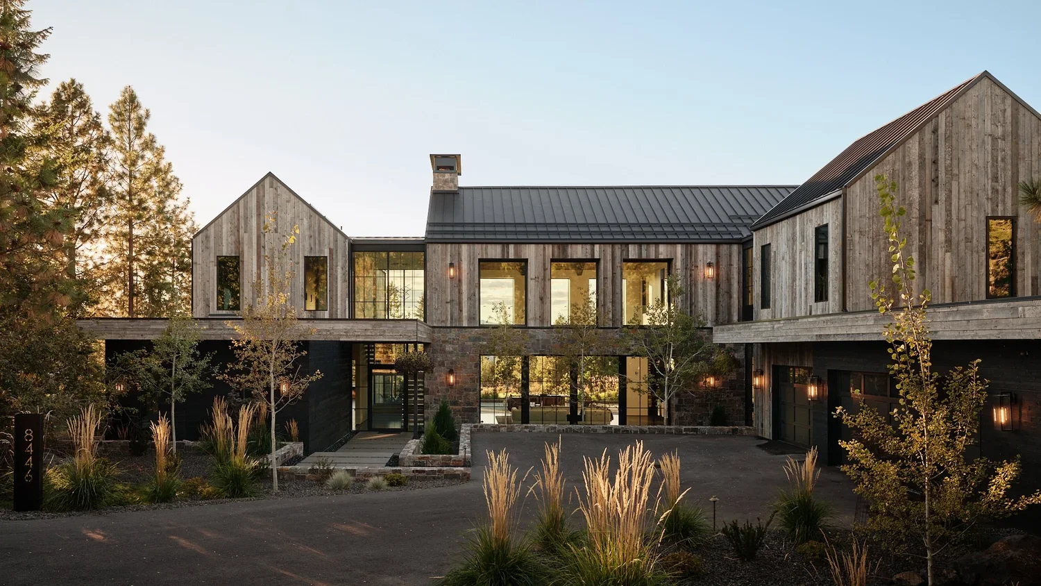 Modern two-story house with wood siding and large windows, surrounded by trees and landscaped yard, during sunset.