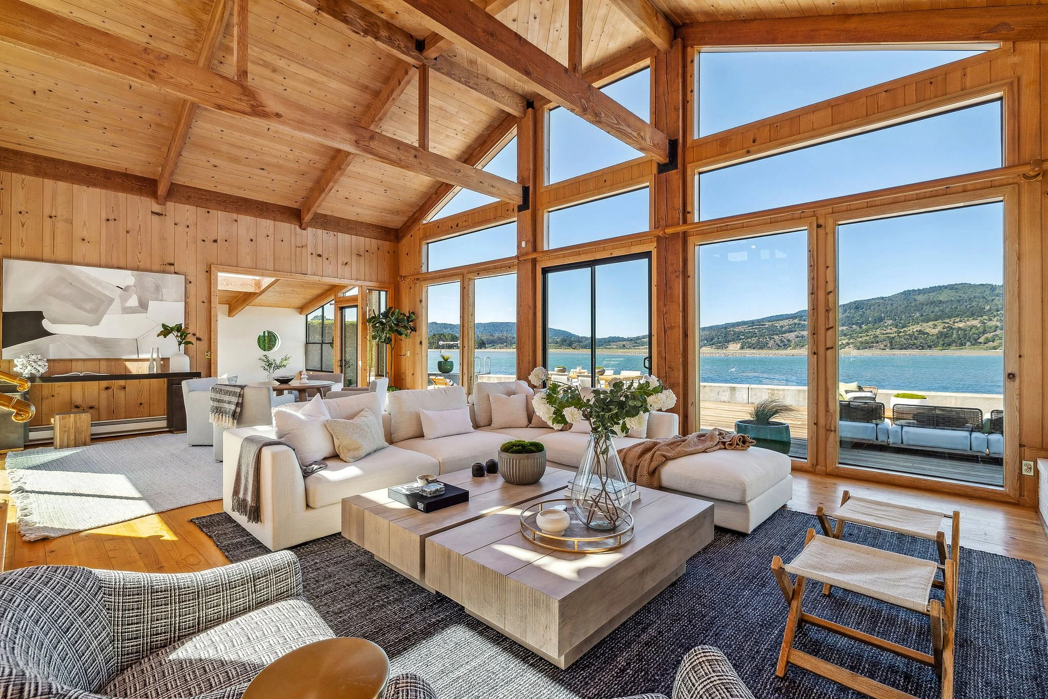 A spacious living room with large glass windows overlooking a lake and mountains, featuring a white sofa, patterned armchair, wooden coffee table, and various plants and decorations, with wooden walls and ceiling.