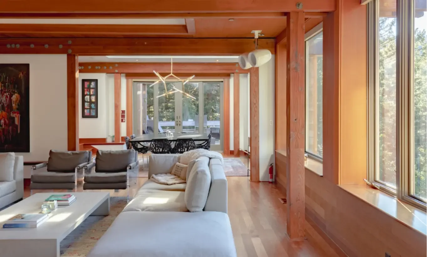 Living room with large windows, wooden beams, modern white sofas, black chairs, and a dining table with a distinctive light fixture