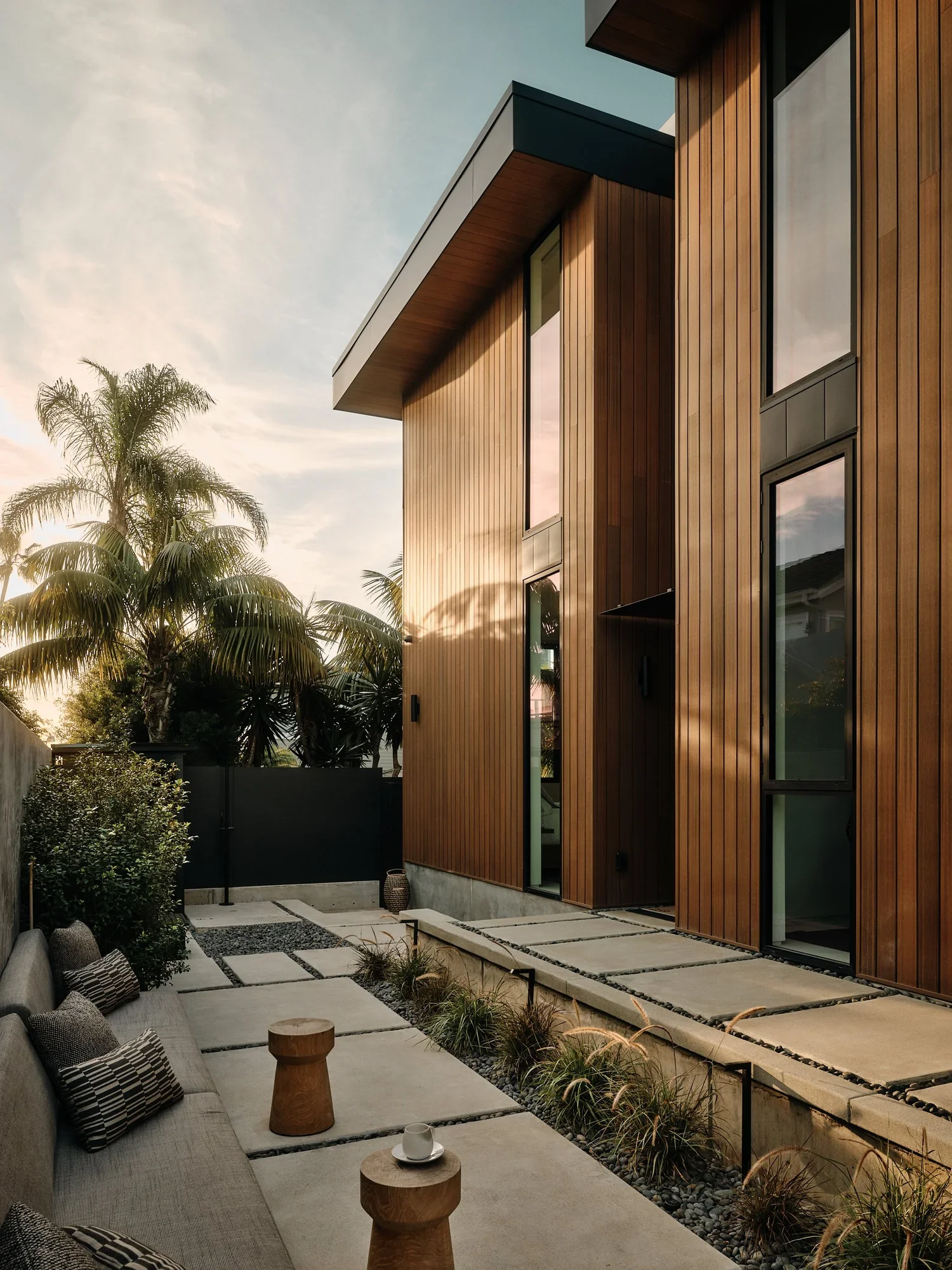 Modern house with wooden exterior panels, large windows, and a backyard patio with outdoor seating, plants, and decorative rocks at sunset.
