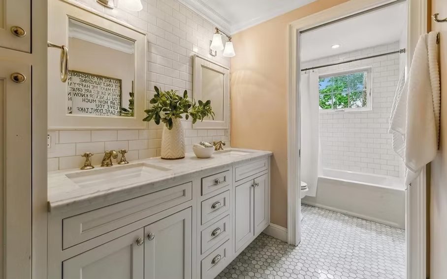 Bright bathroom with white vanity, gold fixtures, mirror, and green plant, connected to a shower area with a small window.