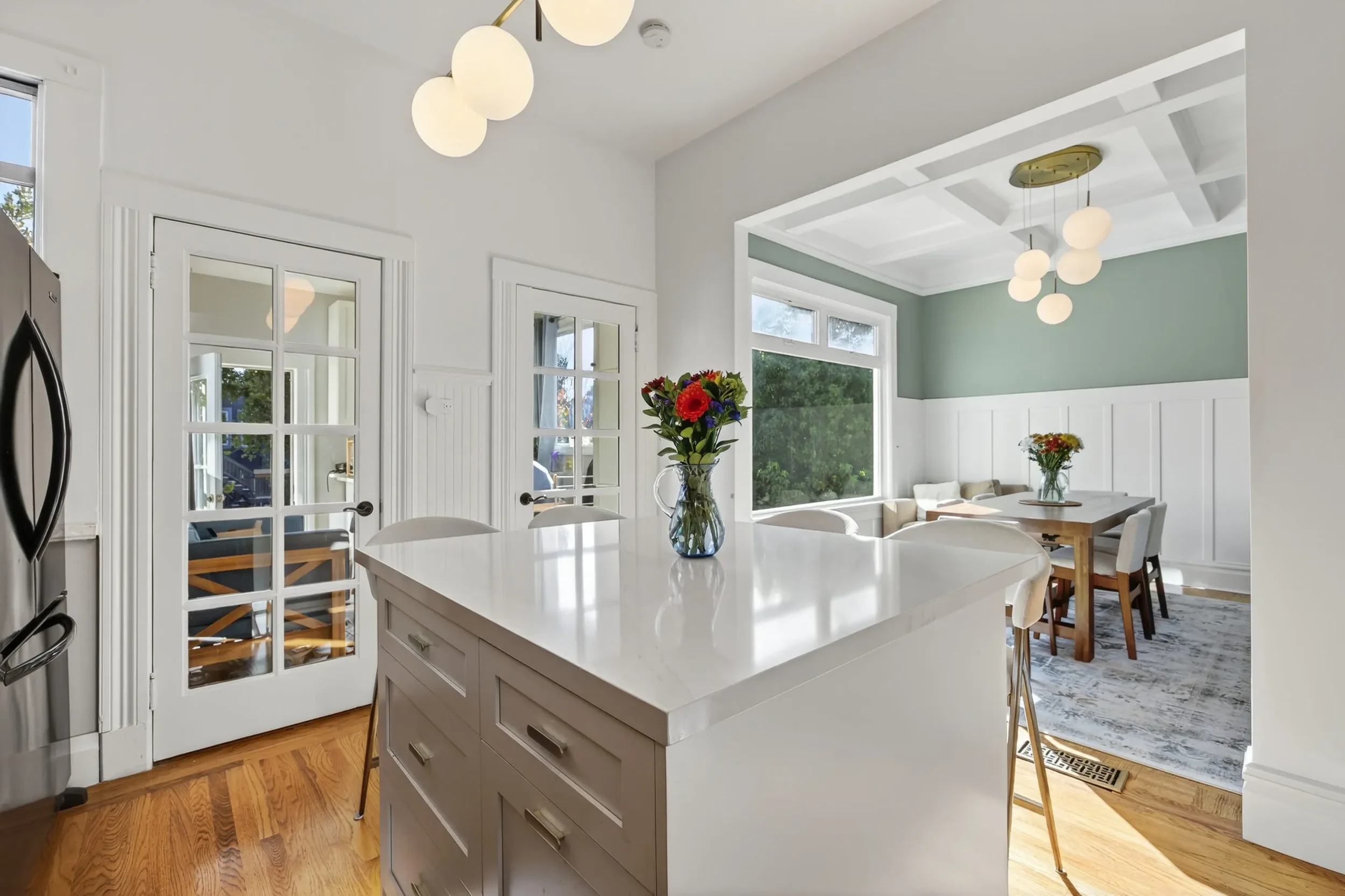 Bright, modern kitchen with white cabinets and island, wooden flooring, and large windows providing natural light. A vase with flowers sits on the island, and a dining area with a table and chairs is visible in the background.