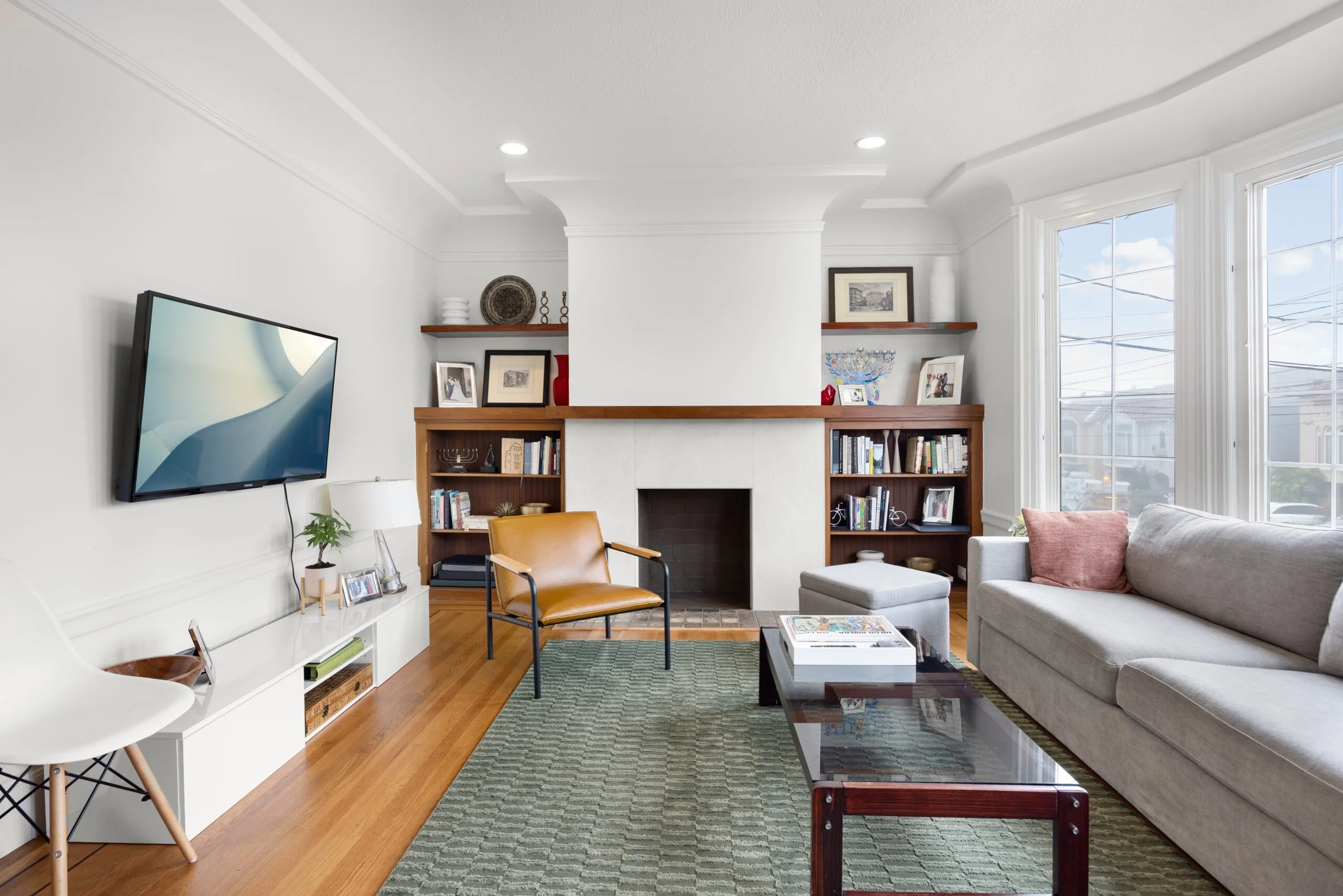 Living room with white walls, wooden bookshelves, a gray sofa, a leather armchair, a coffee table, a large window, and a mounted flat-screen TV.