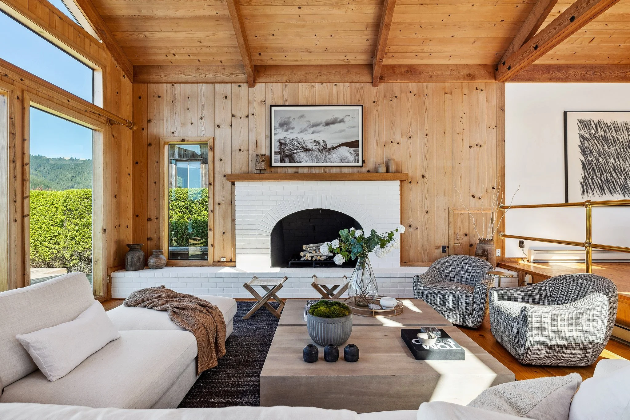 Living room with wood-paneled walls, large windows, a white brick fireplace with a black and white horse photograph above, and cozy seating including a white sofa and patterned chairs, decorated with plants and decorative objects.