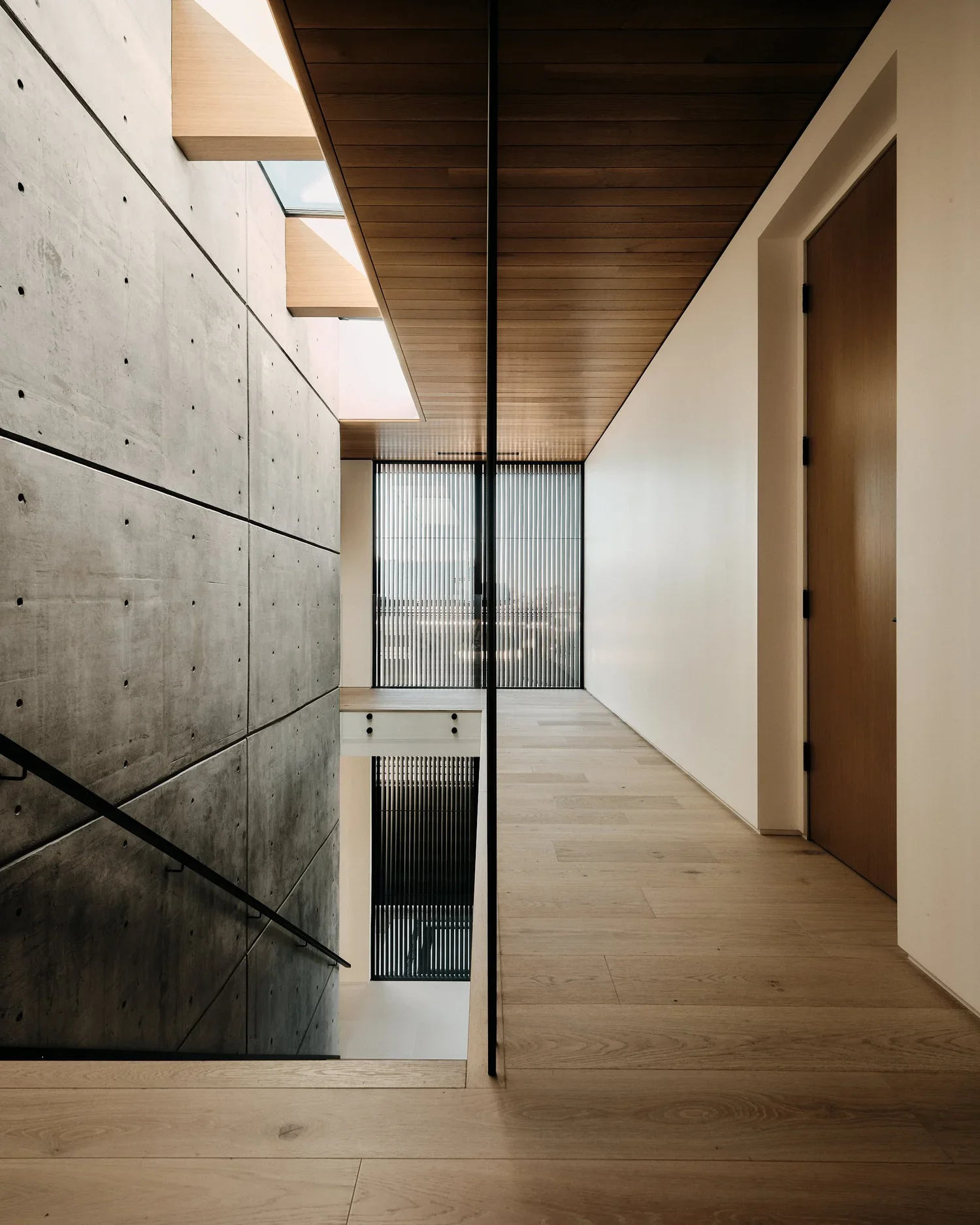 Interior view of a modern building with concrete stairs, wooden accents, a hallway with wooden flooring, white walls, and large windows with vertical blinds.