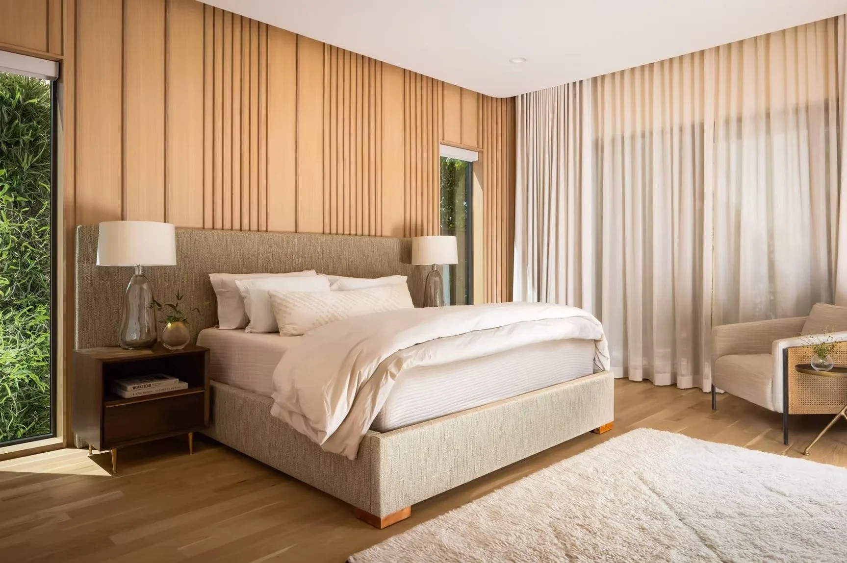 Modern bedroom with a large bed, nightstands with lamps, and a sitting area, featuring wooden accent wall, large window with white curtains, and a sliding glass door overlooking greenery.