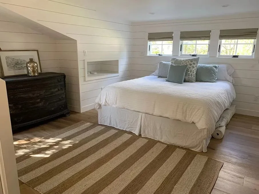 A cozy bedroom with white shiplap walls, a large bed with white bedding, decorative pillows, and three small windows above the bed. There is a dark wooden dresser with decorative items, and a striped rug on the wooden floor.