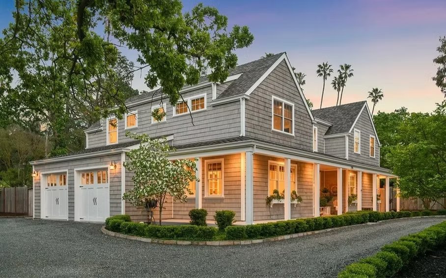 A two-story gray house with a covered porch, surrounded by landscaped greenery and tall trees at sunset.