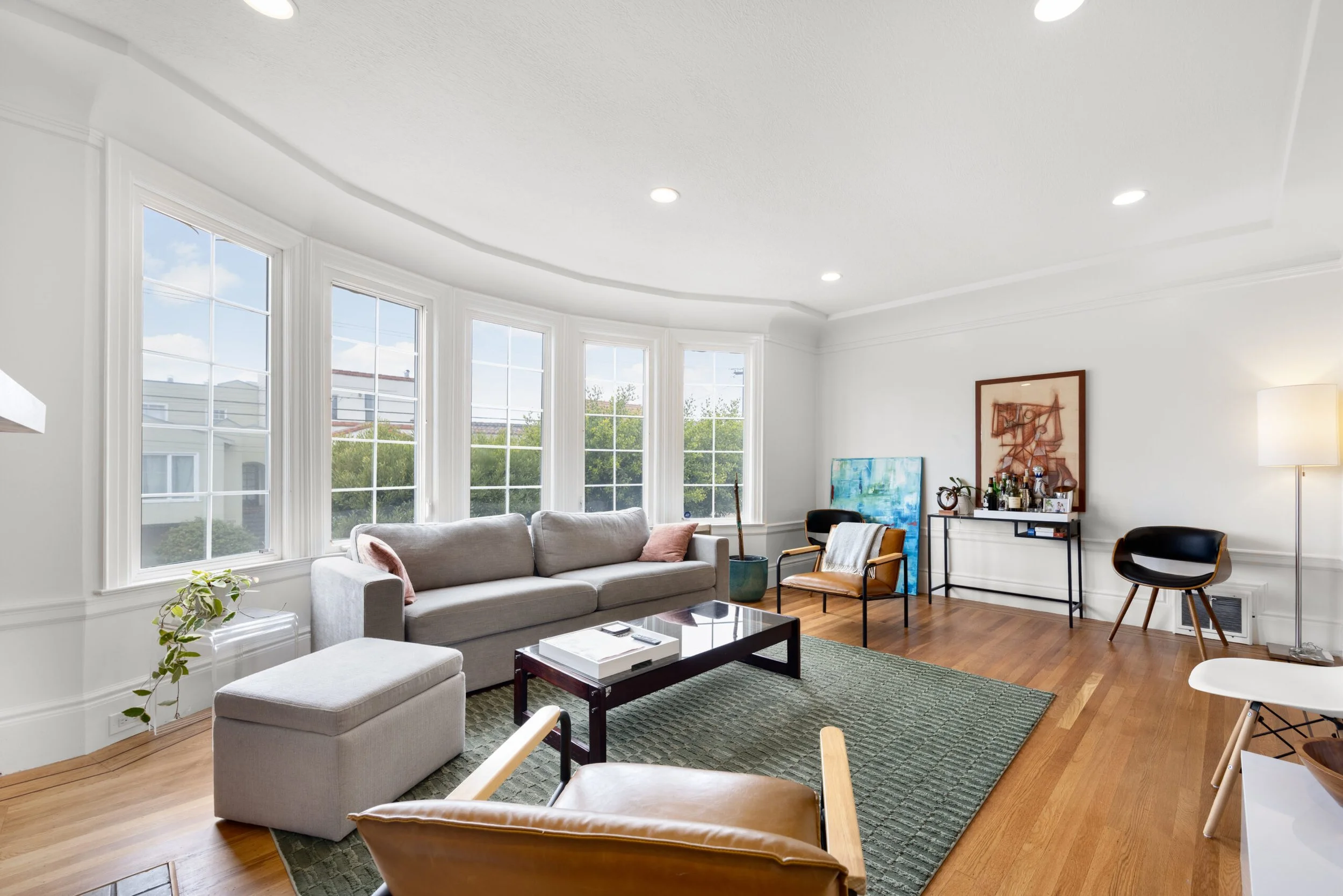 Living room with large windows, white walls, hardwood flooring, modern furniture including a sofa, chairs, and a coffee table, decorated with paintings, plants, and art pieces, illuminated by natural light and ceiling lights.