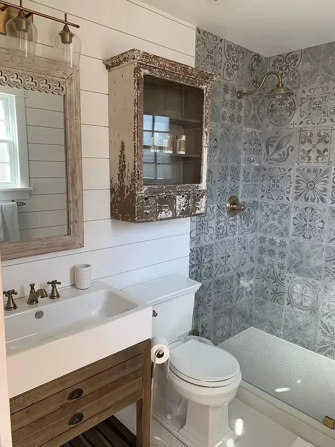 Bathroom with a white sink, wooden cabinet, and a vintage distressed wall cabinet with glass door. Shower area with patterned gray tiles and a brass showerhead. White toilet with a toilet paper roll. Mirror with a wooden frame and a small window with