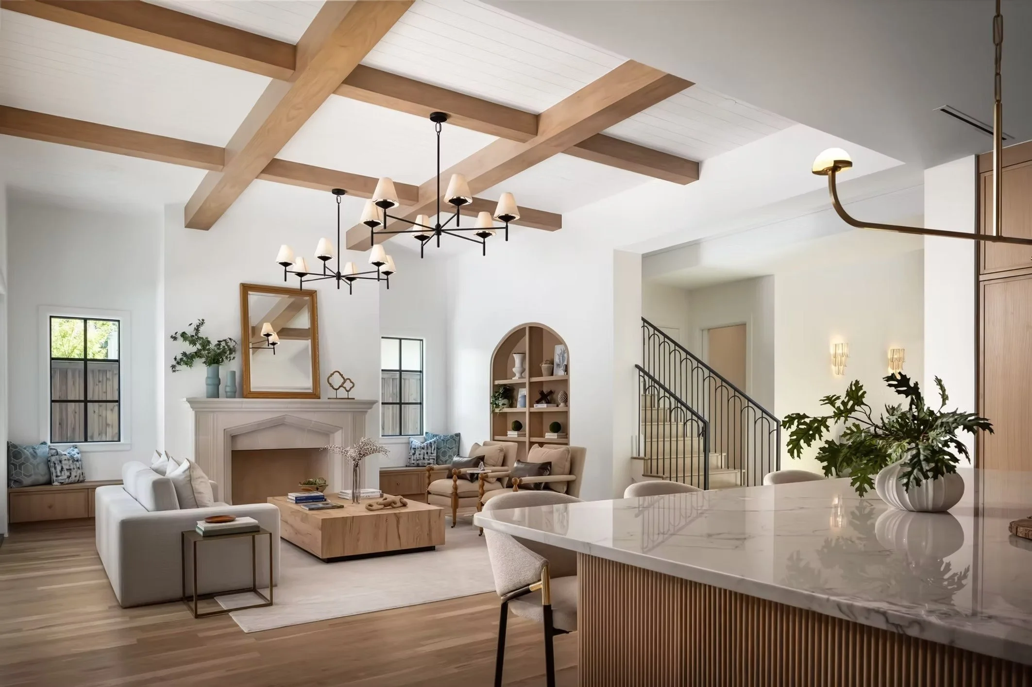 Open concept living room and dining area with wood beam ceiling, modern chandeliers, and a white fireplace, decorated with plants, mirrors, and cozy furniture.