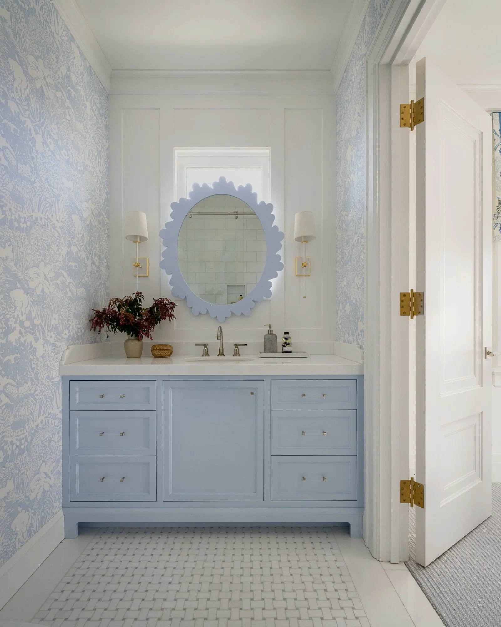 Bathroom vanity with a round mirror, white countertop, blue cabinet with gold handles, wall-mounted lights, a vase with flowers, and a window reflected in the mirror.