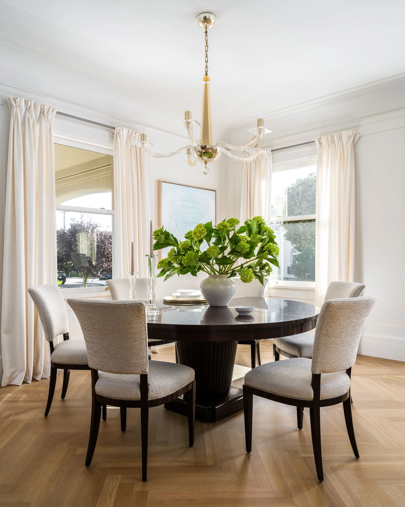 A dining room with a round black table, six beige upholstered chairs, a chandelier with glass arms, a white vase with green hydrangeas, and a window with cream curtains.