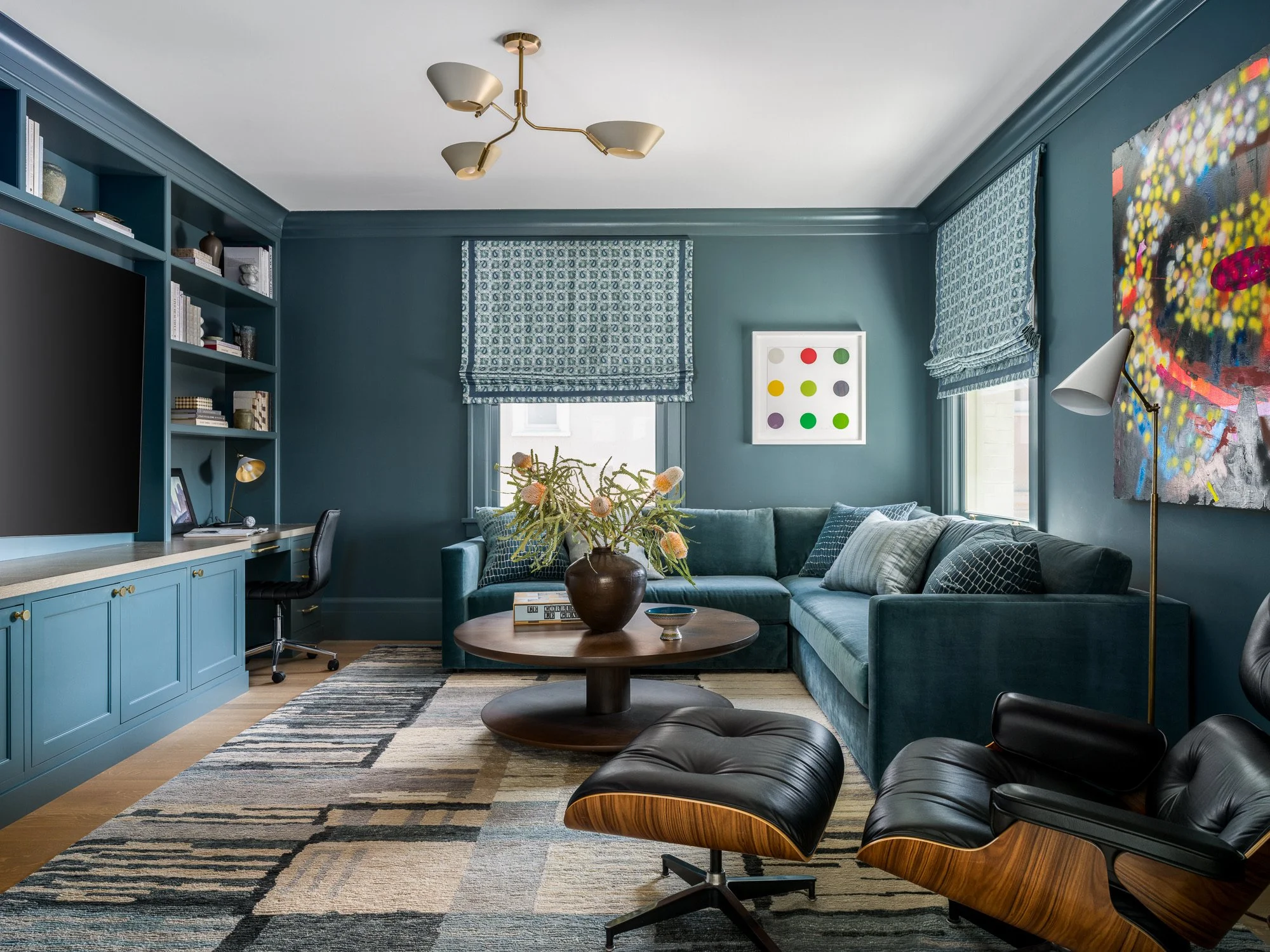 Living room with blue walls, blue curtains, a teal sectional sofa, black lounge chairs with wooden accents, a round wooden coffee table, wall art, bookshelf, and a patterned rug.