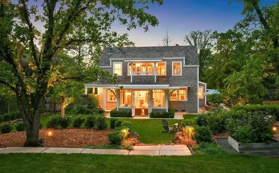 A two-story house with a porch, large windows, and a garden in the evening, with warm lights inside