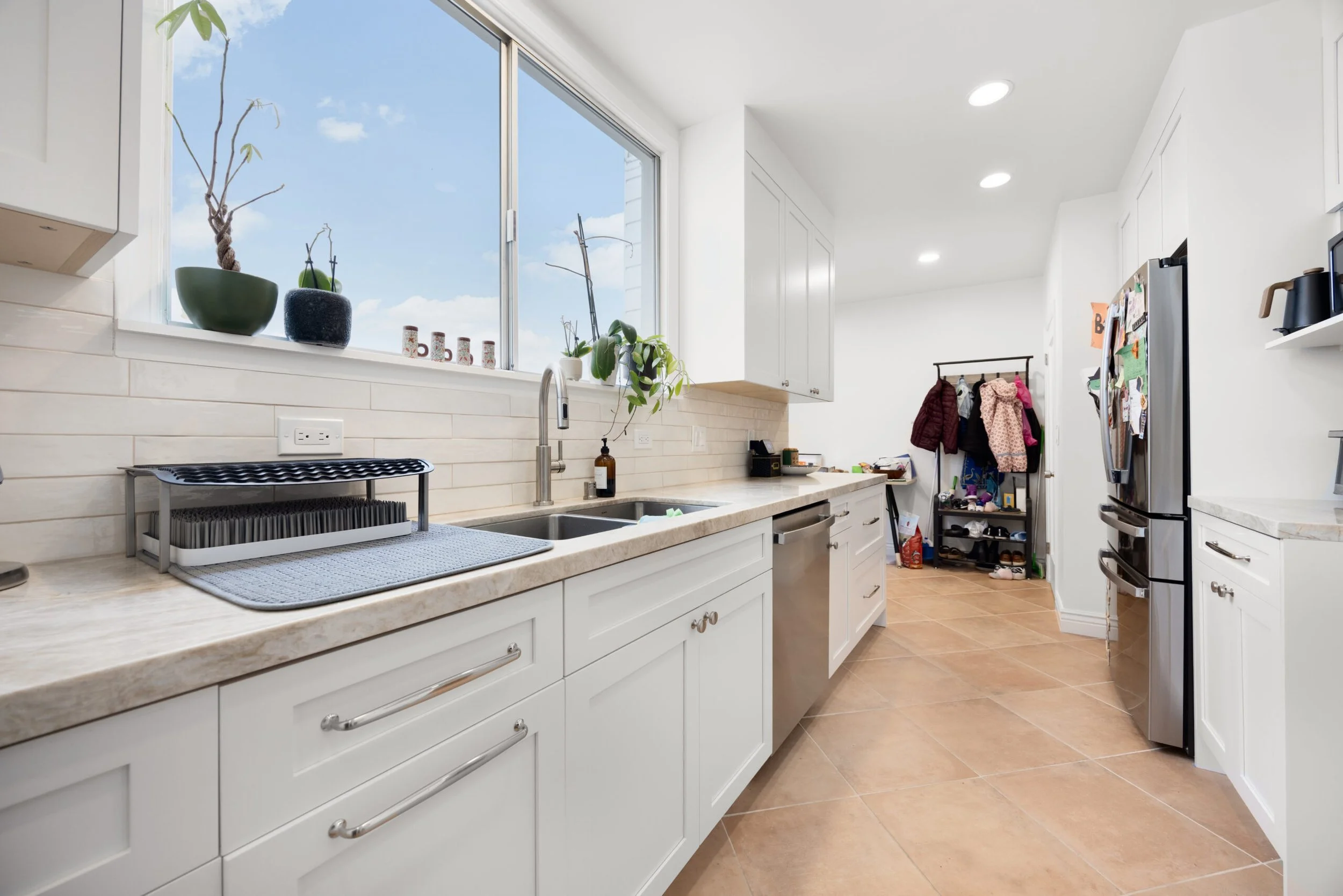 A bright kitchen with white cabinets, a beige marble countertop, and a large window with blue sky and clouds visible. The kitchen features a stainless steel sink, a dish drying rack, and a few potted plants on the windowsill. In the background, there