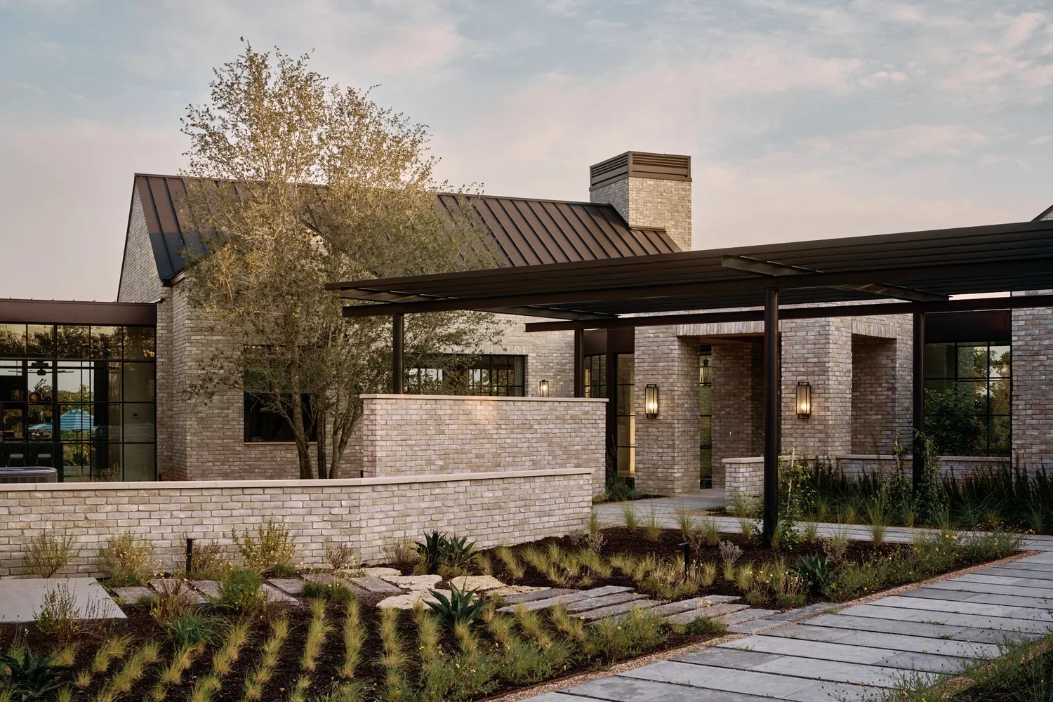Modern house with brick exterior, large windows, and a landscaped pathway with plants and a tree in front.