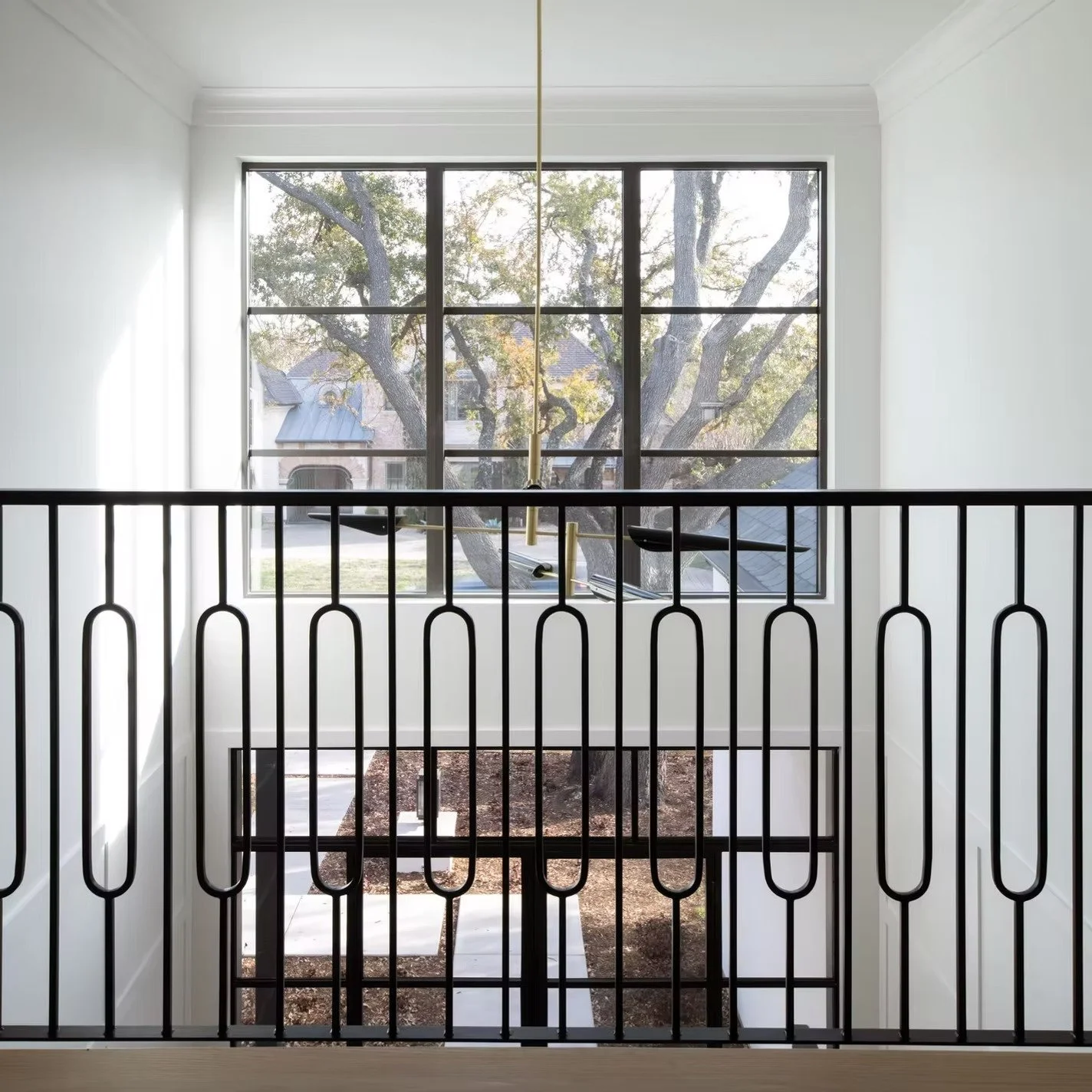 View from a second-floor balcony overlooking a porch and street with trees and houses, black metal railing in foreground, large window with divided panes in the background, interior painted in white.