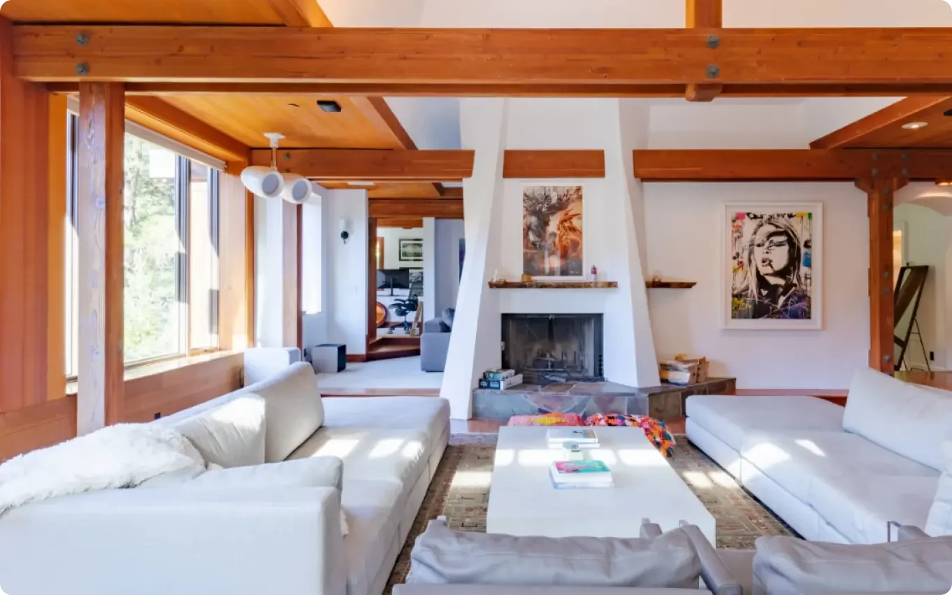 Living room with white sofas, a fireplace, wooden beams, large windows, and contemporary artwork.
