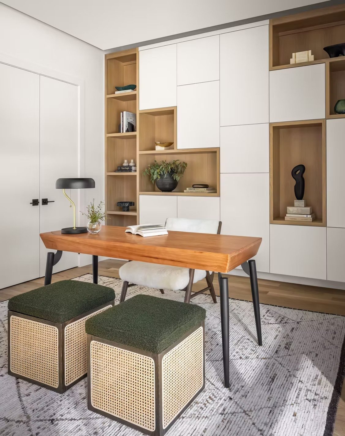 Interior of a modern home office with a wooden desk, two green upholstered stools, a white chair, and a black desk lamp. A decorative shelf wall with books, plants, and art adorns the background.